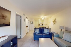 Ein Wohnzimmer mit blauem Sofa, einem weißen Couchtisch und einem grauen Sofa. Im Hintergrund ist eine Essecke mit Tisch und Stühlen zu sehen.
