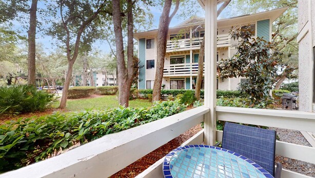 A covered outdoor area with a tiled pool table, a white railing, and a building with multiple balconies in the background. - Coastal Retreat with Patio, Shared Pools, Tennis & Playground - Walk to Beach (Hilton Head)