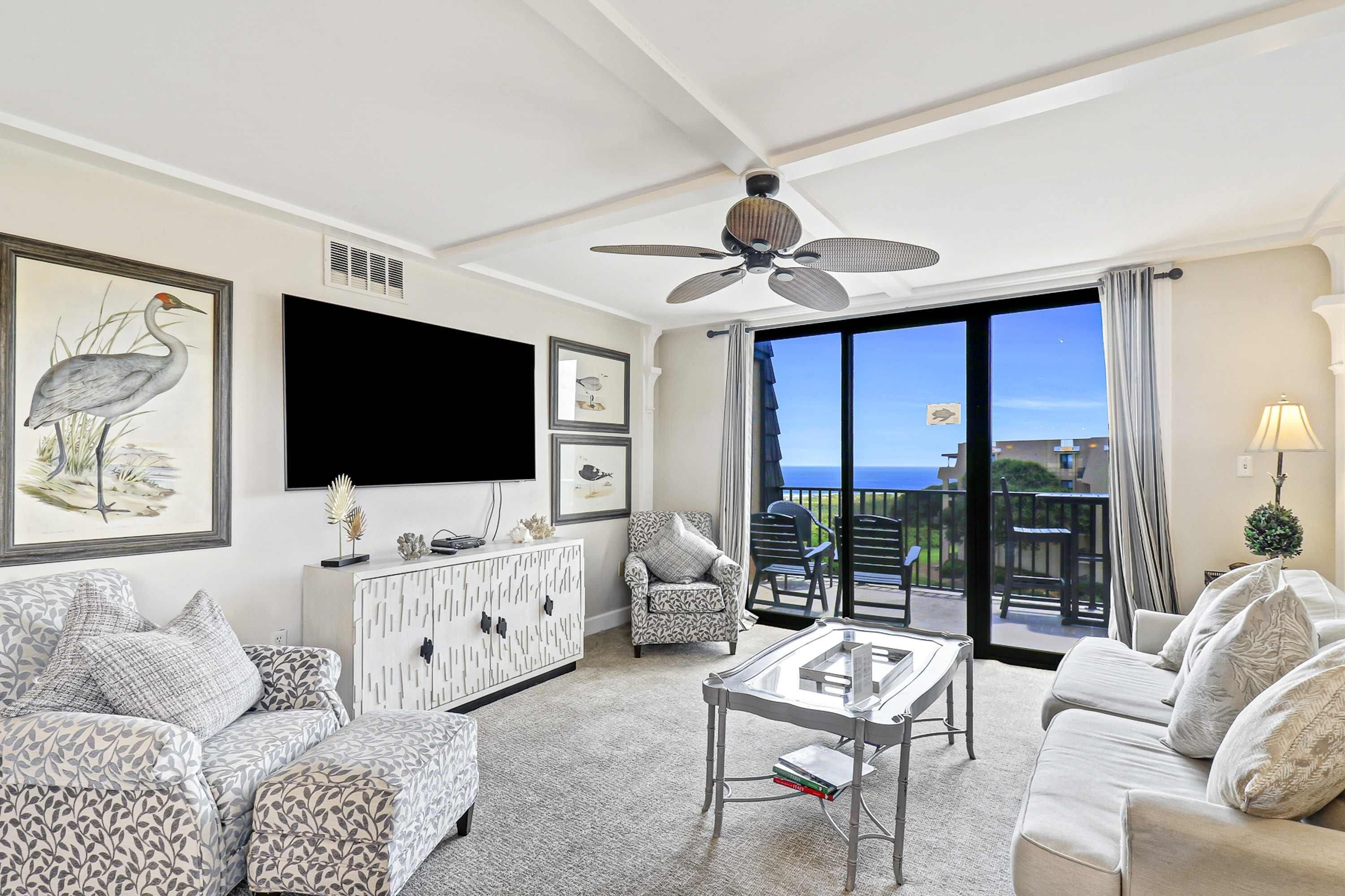 A living room with a sofa, armchair, coffee table, ceiling fan, TV, artwork, and a view through a sliding glass door.