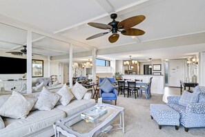Ein geräumiger Wohnbereich mit Sofa, Sessel und Hocker, einem Esstisch mit Stühlen, Deckenventilator und einer Küche im Hintergrund.