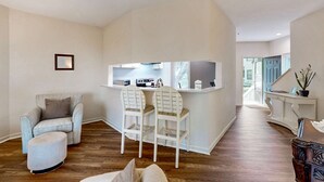 Ein Wohnzimmer mit Sofa, Hocker und einem Essbereich mit Stühlen sowie einer Küchenzeile.