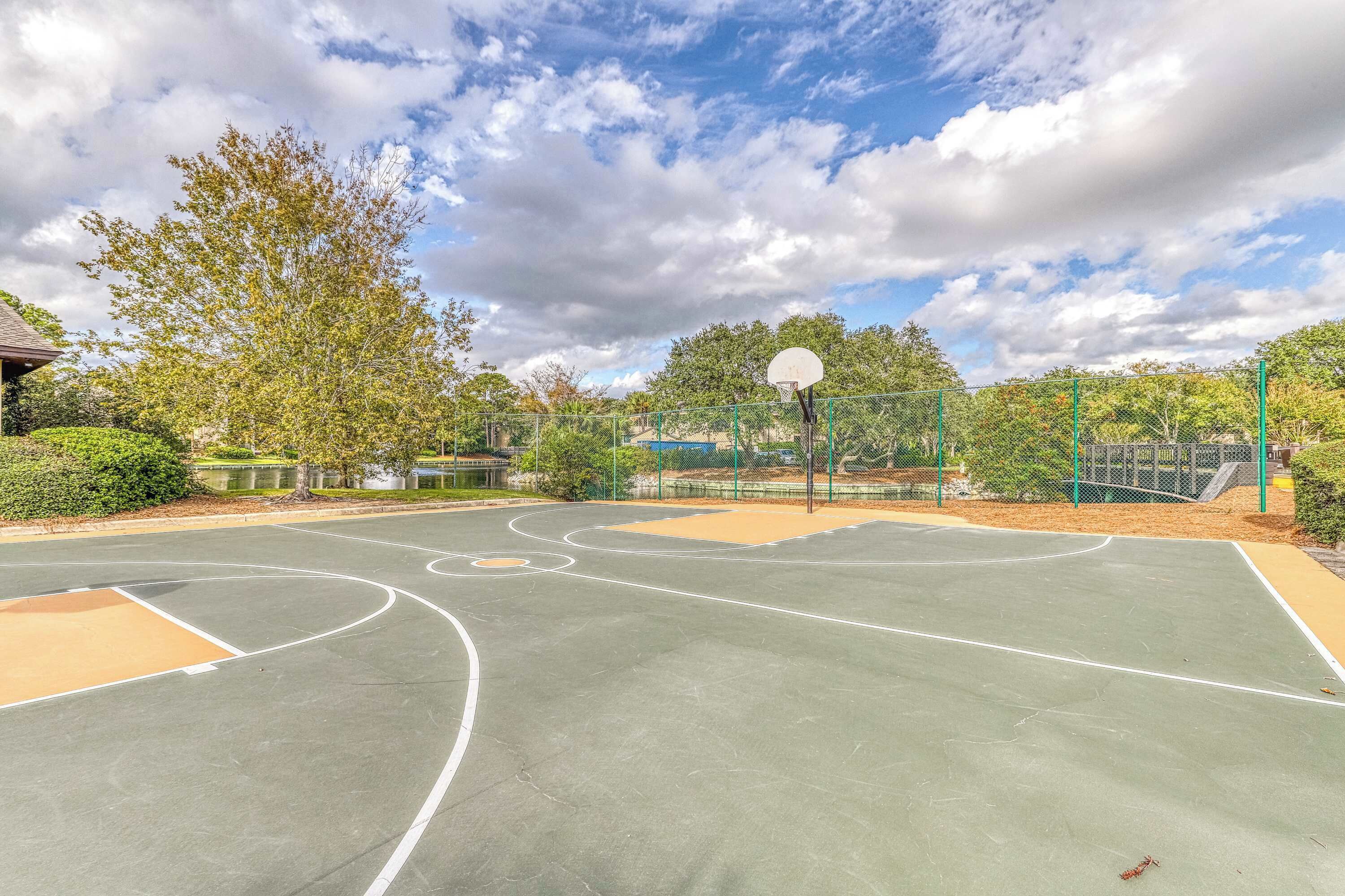 Una cancha de baloncesto al aire libre con aro, rodeada de árboles y una valla.