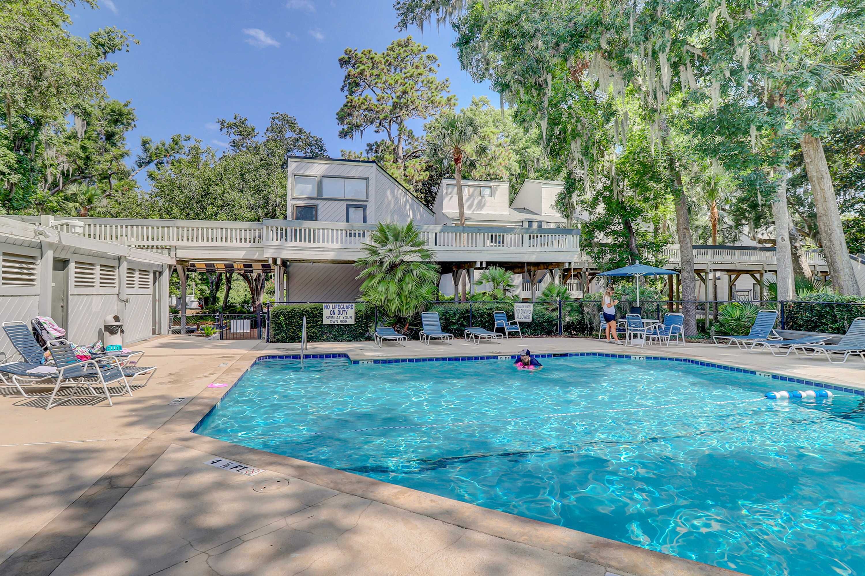 A swimming pool with lounge chairs, a covered deck, and a two-story building with a balcony.