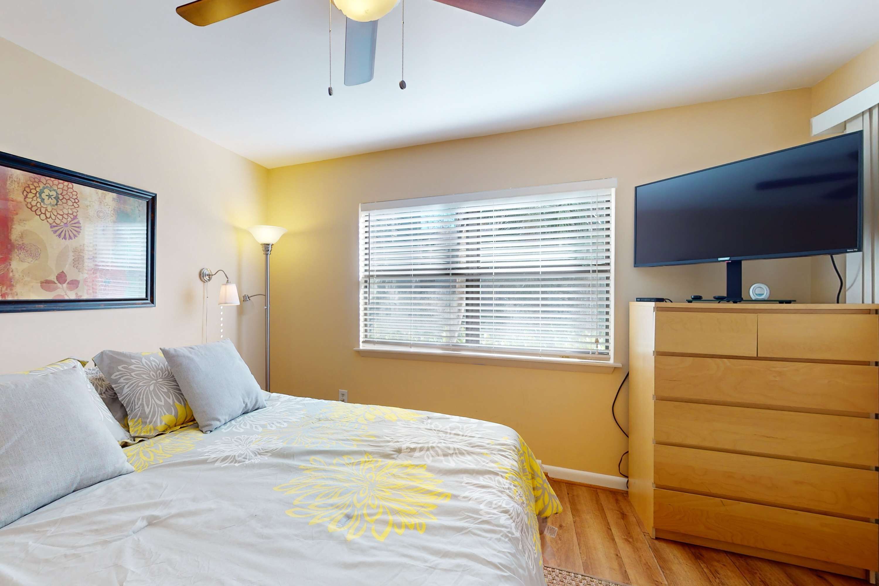 A bedroom with a bed, a television on a dresser, a window with blinds, and a ceiling fan.