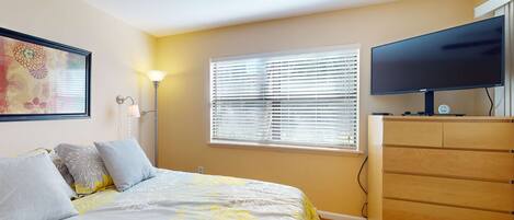 Un dormitorio con cama, televisor sobre una cómoda, ventana con persianas y ventilador de techo.