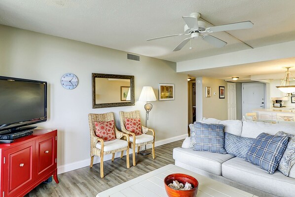 A living room with a television, two chairs, a sofa, a ceiling fan, and a dining area in the background. - Oceanfront condo with WiFi, pool, hot tub & beach access (Hilton Head)