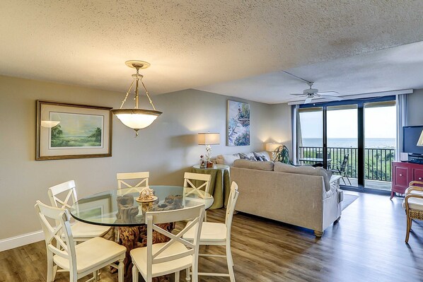 A living room with a glass dining table, white chairs, a sofa, a TV, and a balcony with a view. - Oceanfront condo with WiFi, pool, hot tub & beach access (Hilton Head)