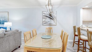 Een eethoek met een tafel met glazen blad, houten stoelen, een moderne kroonluchter en een schilderij aan de muur.
