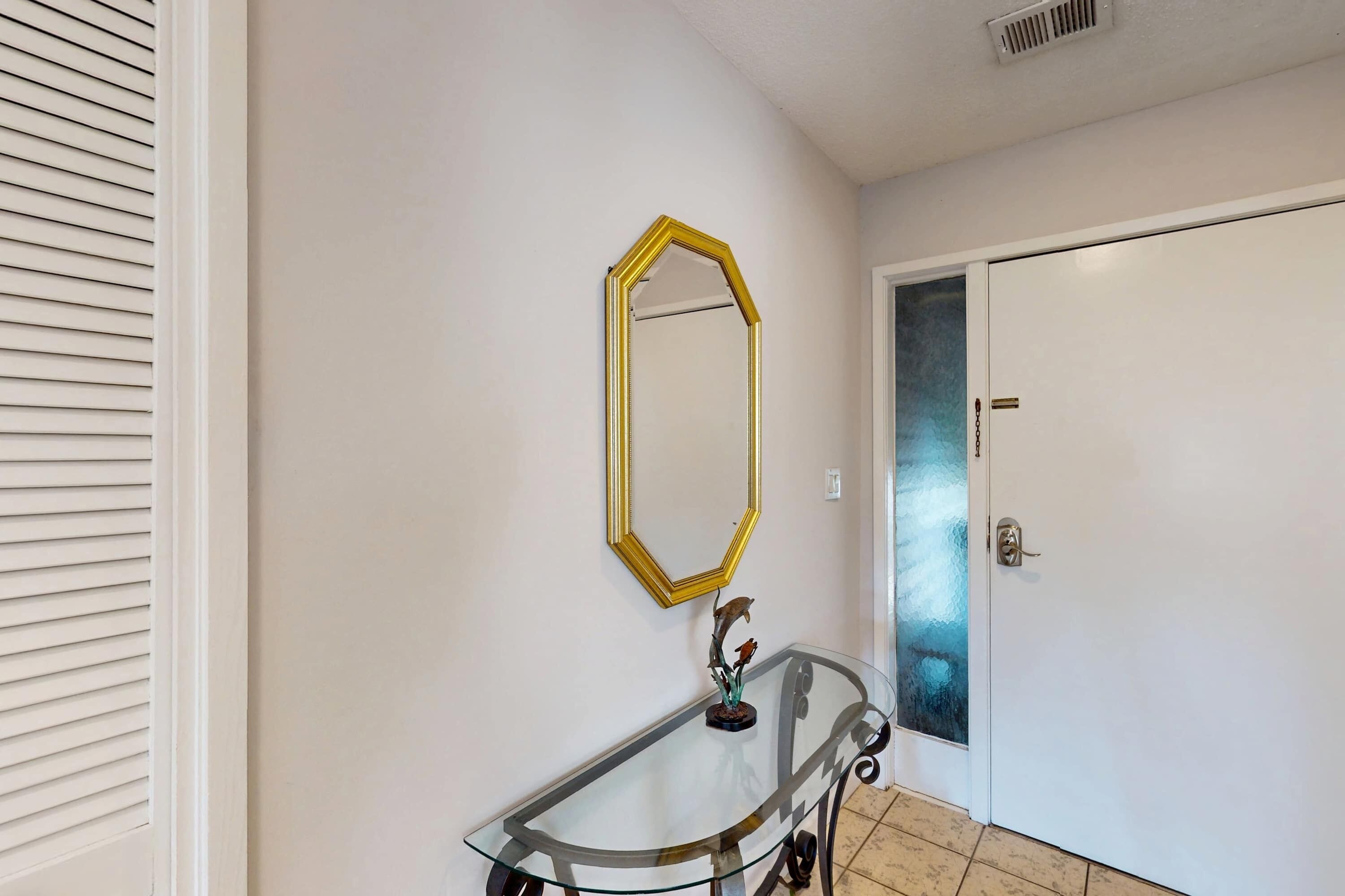 A hallway with a glass-top console table, a gold-framed mirror, and a white door with a frosted glass panel.