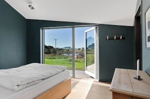 A bedroom with a bed, a wooden nightstand, a large sliding glass door leading to a balcony with a view of a green landscape and mountains.