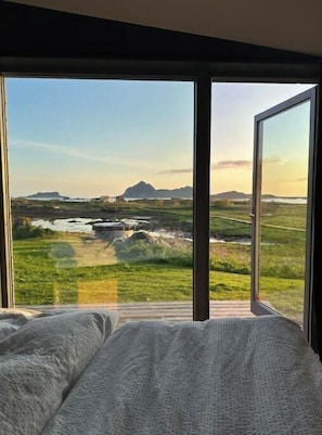 A bedroom with a large window, a bed, and a view of a landscape with mountains and a body of water.