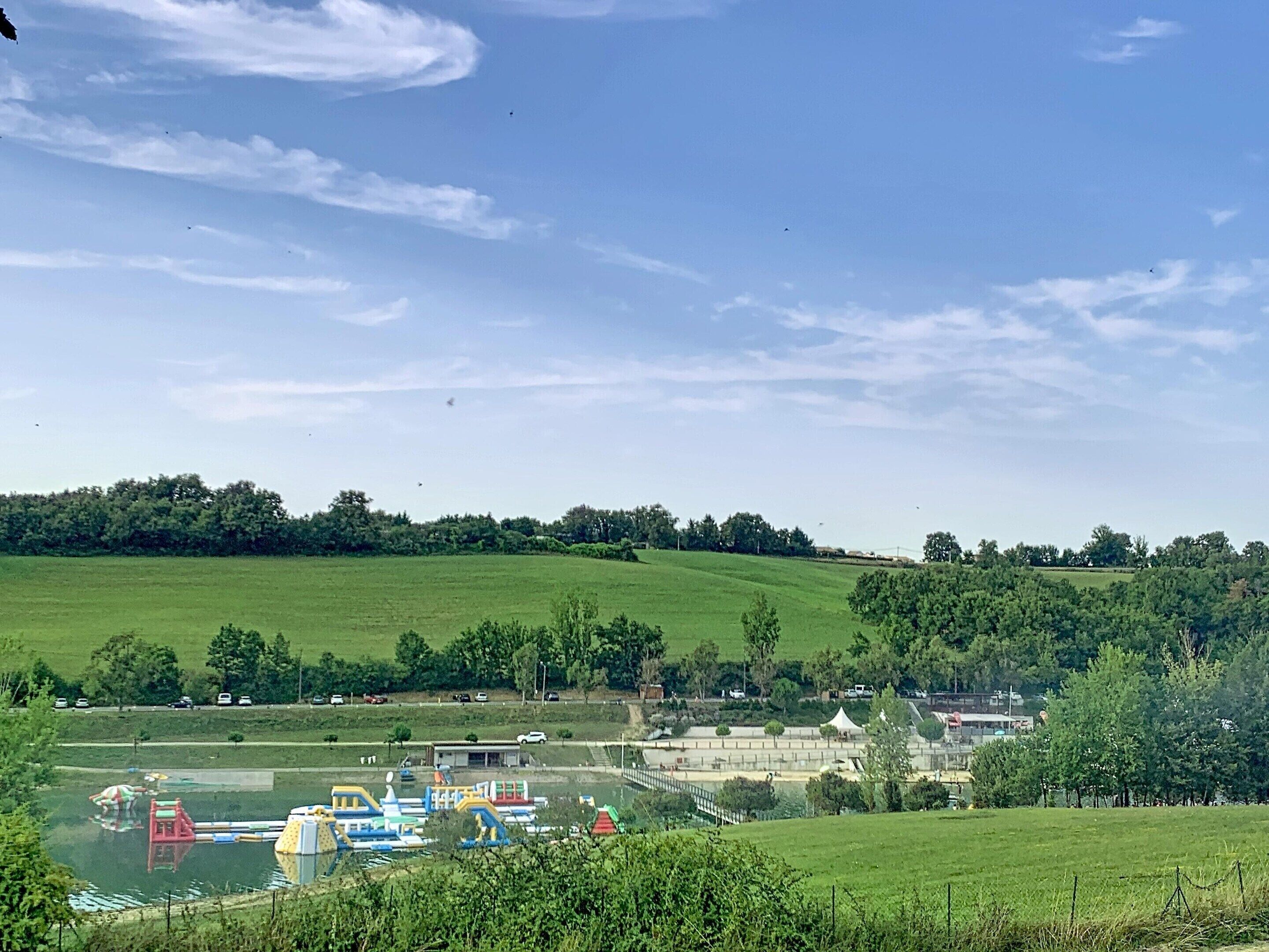 Paesaggio rurale con un prato verde, un piccolo edificio e una distesa d'acqua con strutture gonfiabili.