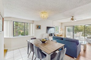 A modern living room with a dining table, sofa, TV, and large windows. - Inviting family-friendly condo with balcony, central AC, & outdoor pool (Hilton Head)