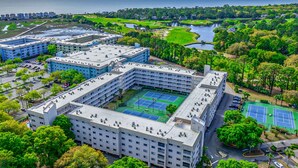 Ett hotell med flera våningar, en tennisbana och omgivet av grönska och en vattenyta.