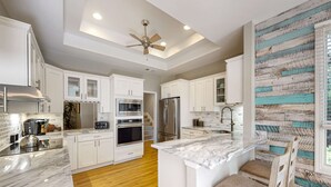A modern kitchen with white cabinetry, a marble countertop, a ceiling fan, and a rustic wall. - Bright & Expansive Beach Retreat with Private Pool (Hilton Head)