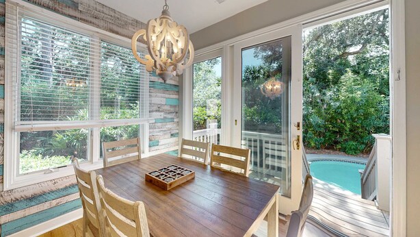 A dining area with a wooden table, chairs, a chandelier, and large windows with a view of a swimming pool and greenery. - Bright & Expansive Beach Retreat with Private Pool (Hilton Head)