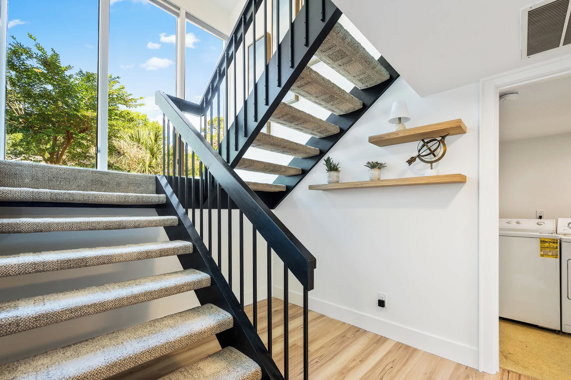 A modern staircase with a black railing, a kitchen area with appliances, and a view of greenery outside.