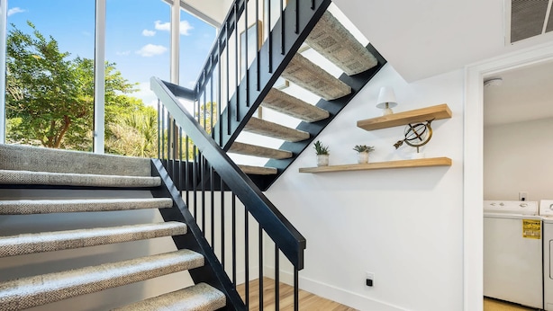 Un escalier moderne avec une rampe noire, un espace cuisine équipé d’appareils électroménagers et une vue sur la verdure à l’extérieur.