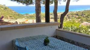 A balcony with a table and chairs, overlooking a coastal landscape with trees and houses. - Panoramic Costa Rei (MURAVERA CA)