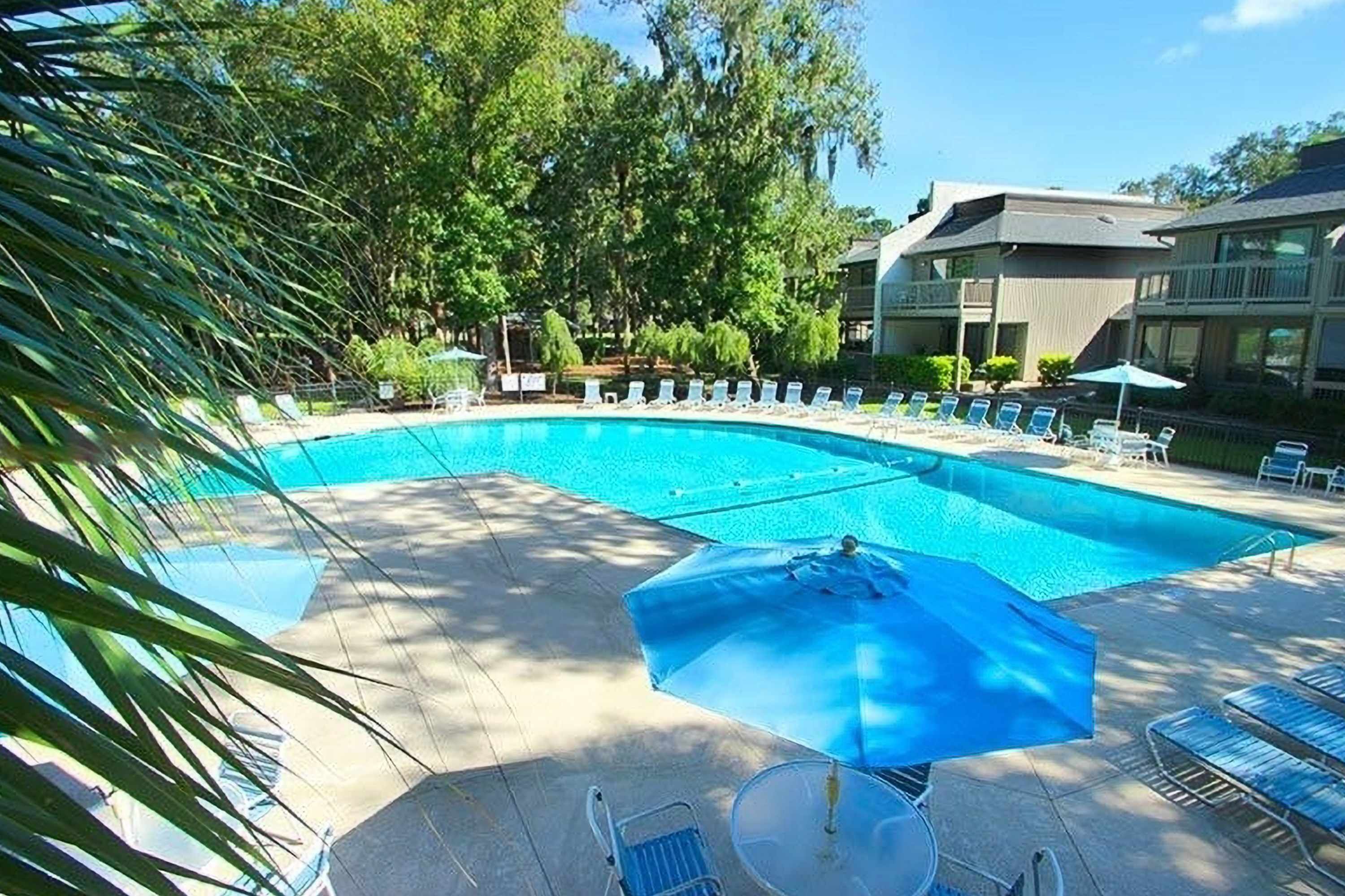 Zona piscina con sedie a sdraio e ombrelloni, circondata da alberi e edifici.