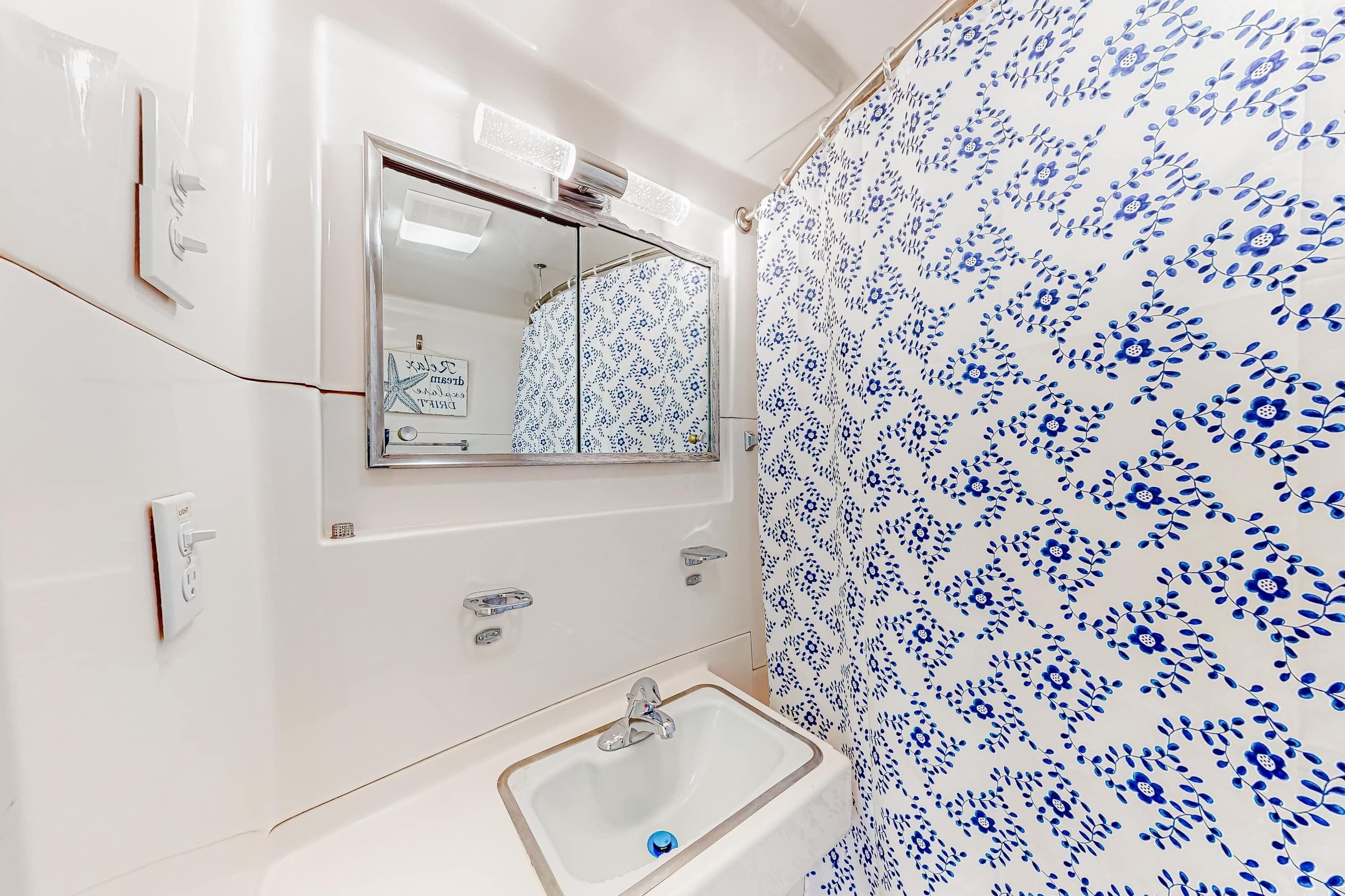Une salle de bain avec un lavabo blanc, un miroir et un rideau de douche orné d’un motif floral bleu.