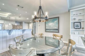 A modern kitchen with a round glass table, wooden chairs, a hanging light fixture, and a white cabinet with decorative items. - Homey condo with ocean views, balcony, pool & easy beach access (Hilton Head)