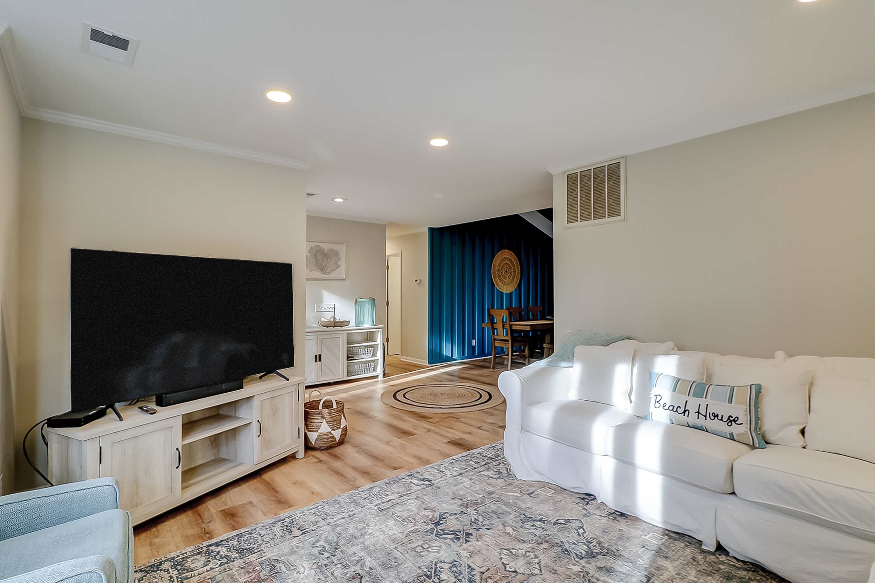 A living room with a flat-screen TV, a white sofa, a patterned rug, and a dining area in the background.