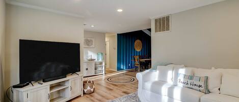 A living room with a flat-screen TV, a white sofa, a patterned rug, and a dining area in the background.