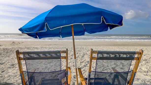 Zwei Liegestühle mit einem blauen Sonnenschirm, ein Holzpfahl und ein Sandstrand mit dem Meer im Hintergrund.
