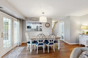 A dining area with a wooden table and white chairs, a kitchen in the background, and a staircase leading to another level. - Renovated Dog Friendly Villa with Patio & Pool  Near Golf & Beach (Hilton Head)