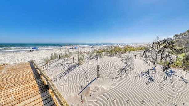 Una playa con un paseo marítimo de madera, dunas arenosas y un cielo azul despejado.