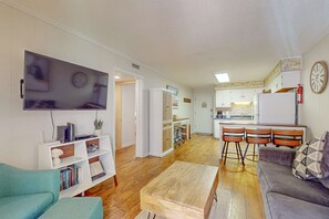 Een woonkamer met een bank, een salontafel, een televisie en een boekenkast. Er is een keuken met een koelkast, een spoelbak en barstoelen.
