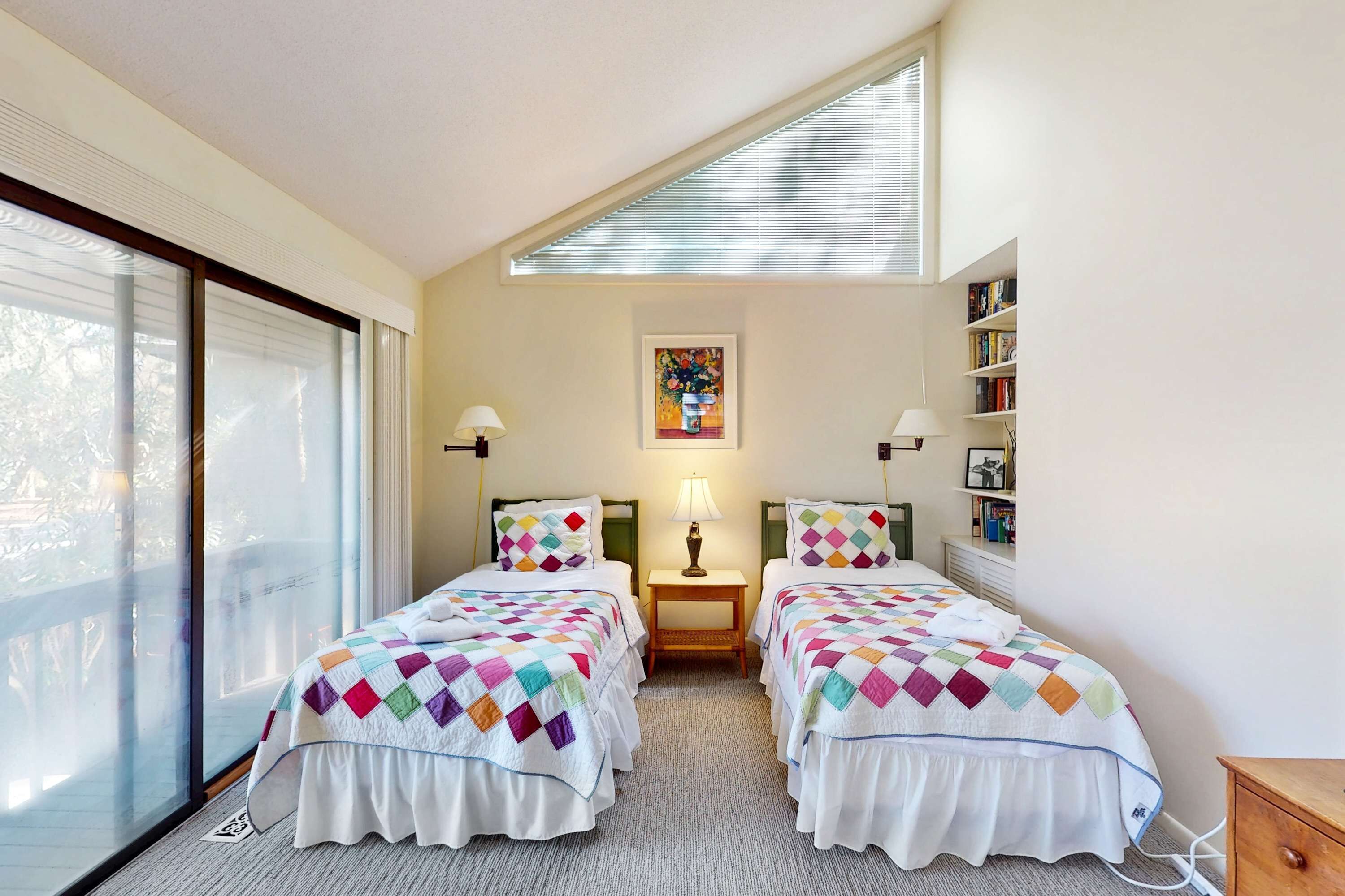 A bedroom with two beds, a window, a lamp, a bookshelf, and a painting on the wall.