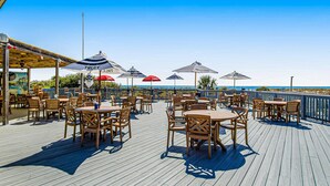 Eine Holzterrasse mit Tischen, Stühlen und Sonnenschirmen, mit Blick aufs Meer.