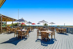 Eine Holzterrasse mit Tischen, Stühlen und Sonnenschirmen, mit Blick aufs Meer.