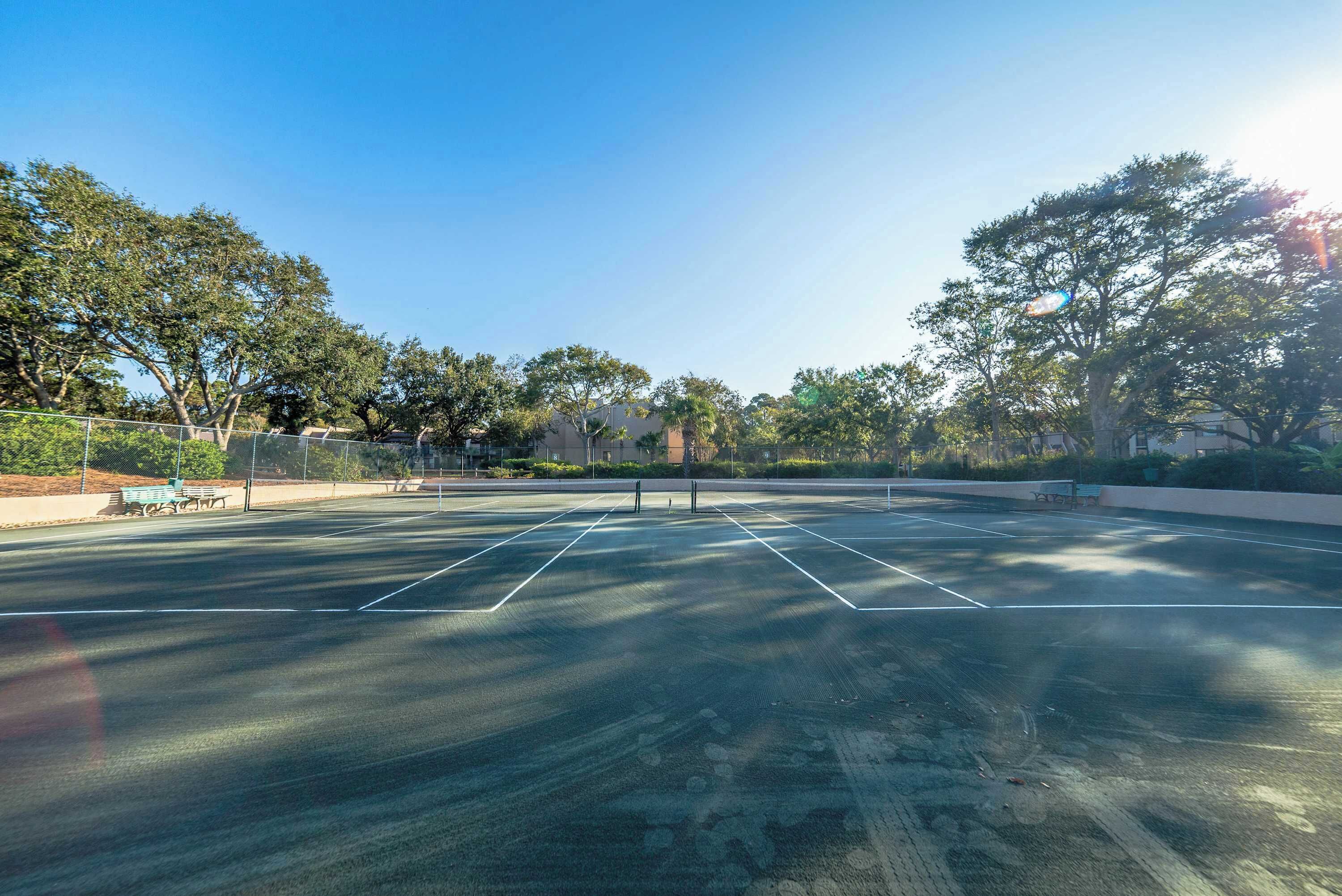 Una cancha de tenis al aire libre con valla, bancos y árboles de fondo.