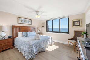 A bedroom with a large window, a bed with a blue patterned bedspread, a wooden nightstand, a chair, and a ceiling fan. - Quiet Waterfront Condo with WiFi - Pools & Hot Tub On-Site (Hilton Head)