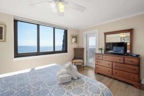A bedroom with a large window, a bed with a patterned bedspread, a wooden dresser with a TV, and a chair. - Quiet Waterfront Condo with WiFi - Pools & Hot Tub On-Site (Hilton Head)
