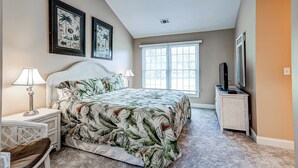 A bedroom with a bed, two wall-mounted picture frames, a bedside table with a lamp, a chair, a TV stand, and a window with blinds. - Intimate, Yet Spacious Villa with Shared Pool and Tennis - Walk to the Beach (Hilton Head)