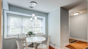 A dining area with a round table, white chairs, a window with blinds, and a staircase leading to an upper level. - Intimate, Yet Spacious Villa with Shared Pool and Tennis - Walk to the Beach (Hilton Head)