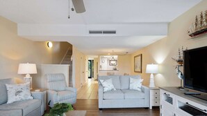Un salon avec un canapé, un fauteuil et un meuble pour la télévision, un escalier et, au fond, une cuisine.