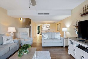 A living room with a sofa, armchair, and TV stand, a staircase, and a kitchen in the background. - Spacious Villa with Shared Pool, Hot tub & Lagoon Views  - Walk to Beach (Hilton Head)