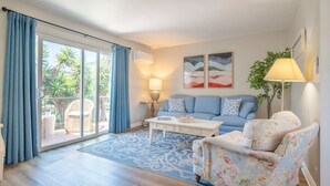 A living room with a sofa, armchair, coffee table, and a view of the outdoors through a sliding door. - Coastal Condo Steps to Beach & Downtown (San Clemente)