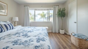 A bedroom with a bed, a window with curtains, a potted plant, a wicker basket, and a framed picture on the wall. - Coastal Condo Steps to Beach & Downtown (San Clemente)