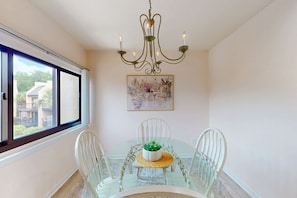 A dining area with a glass table, white chairs, a chandelier, and a painting on the wall. - Elegant Condo with Pool & Hot Tub, Lagoon View & Private Patio (Hilton Head)