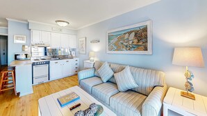 Una sala de estar con un sofá a rayas, una mesa de centro con libros y un control remoto, una cocina con armarios blancos y un cuadro de una escena de playa.