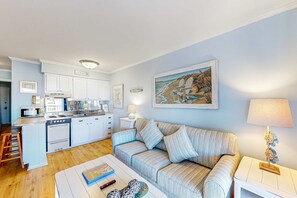 A living room with a striped sofa, a coffee table with books and a remote, a kitchen with white cabinets, and a painting of a beach scene. - 3rd-floor snowbird-friendly condo with pool, tennis, volleyball, gym, & balcony (Hilton Head)