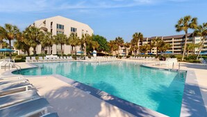 Ein Schwimmbecken mit Liegestühlen, Palmen und einem Hotelgebäude im Hintergrund.