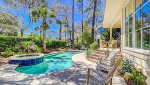 A swimming pool with a curved design, surrounded by lush greenery and tall trees. - Two-story home with pool, balcony, deck, grill, & central AC - snowbirds welcome (Hilton Head)