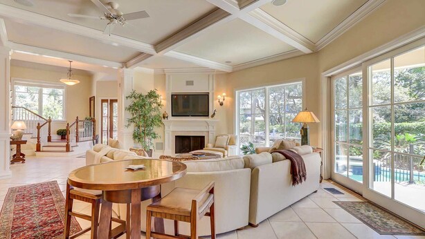 A spacious living room with a fireplace, a television, and a large window overlooking a garden. - Two-story home with pool, balcony, deck, grill, & central AC - snowbirds welcome (Hilton Head)